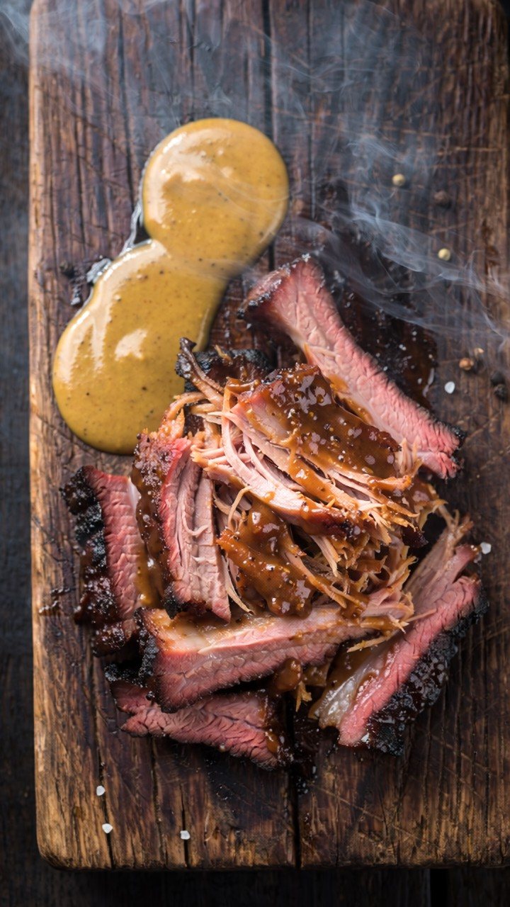 Slices of smoked beef with a dark peppery crust and a pink band just beneath it, some pulled into glossy shreds, on a worn wooden board beside a pool of mustard sauce