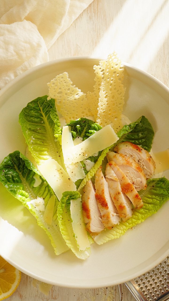 Overhead view of a Caesar salad with whole romaine leaves, shaved Parmesan, lacy Parmesan crisps and sliced grilled chicken in a white bowl.