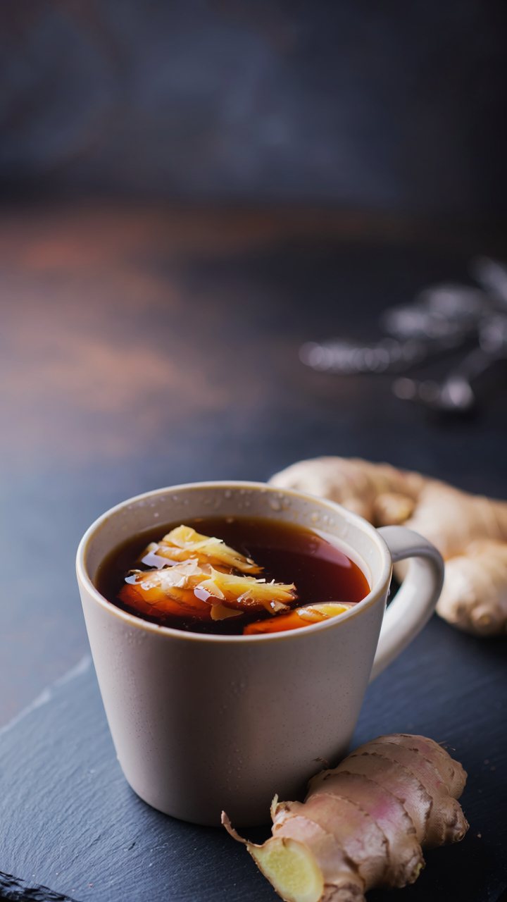 A mug of dark ginger and brown sugar brew with slices of ginger floating in it, knobs of fresh ginger root beside it on dark slate