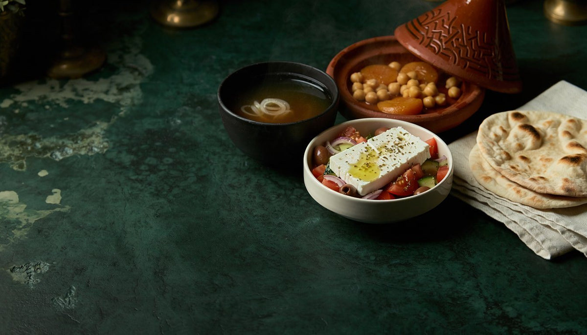 A Greek salad with feta and olive oil, a bowl of clear broth, a clay tagine of chickpeas and apricots and warm flatbreads on a dark green table