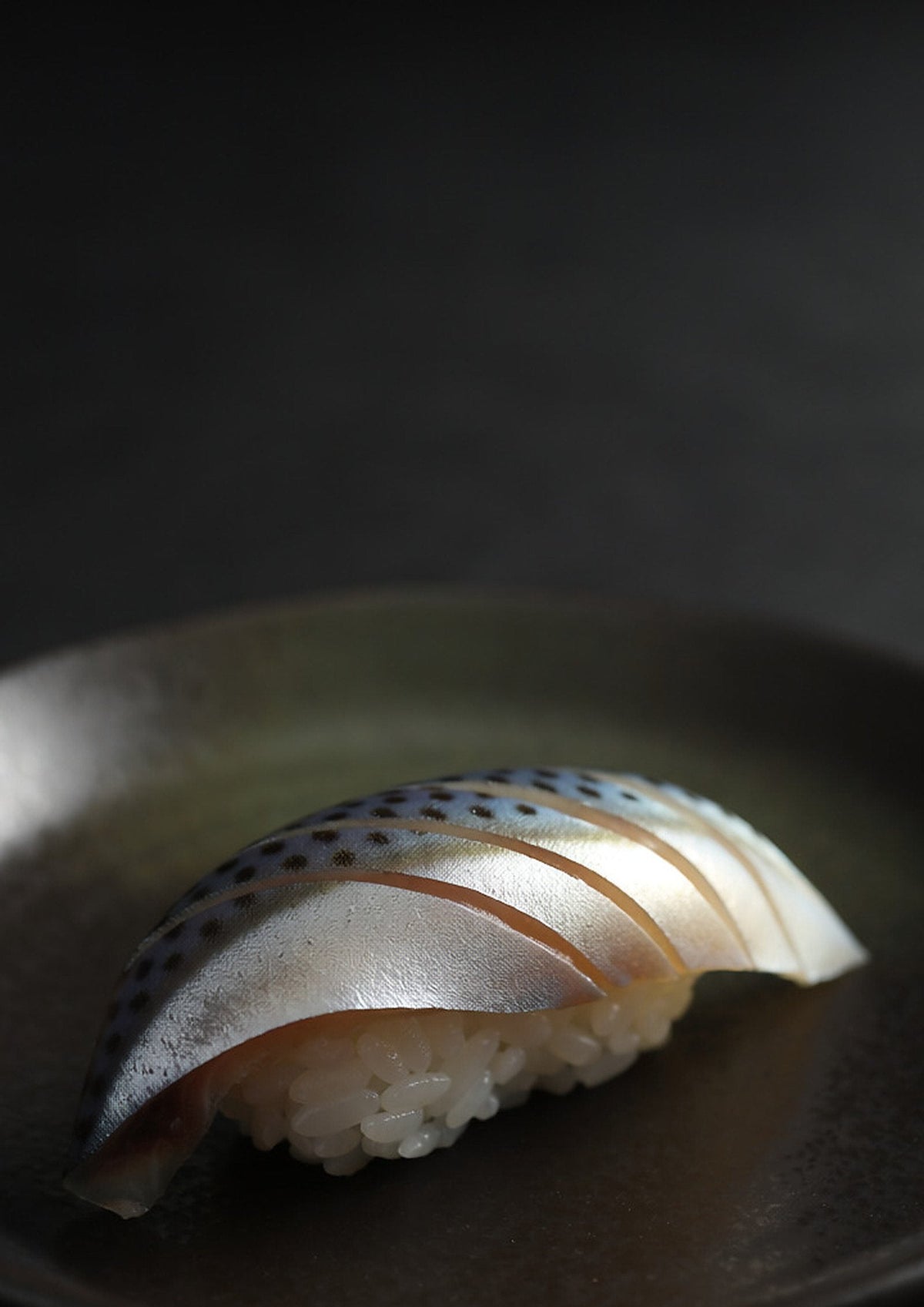 A single piece of kohada nigiri, the spotted silver skin of the cured fish finely scored, on a dark ceramic plate