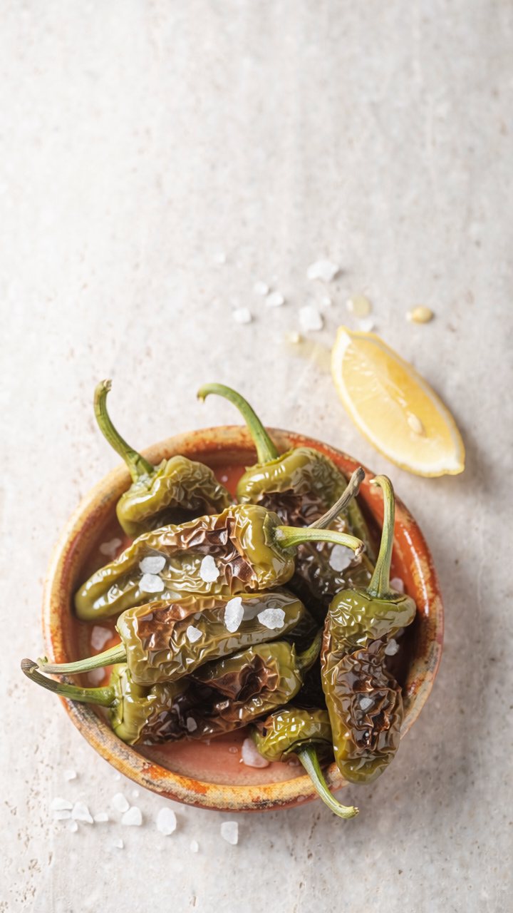 Overhead view of blistered green Padrón peppers scattered with large flakes of sea salt in a rustic terracotta bowl, with a lemon wedge.