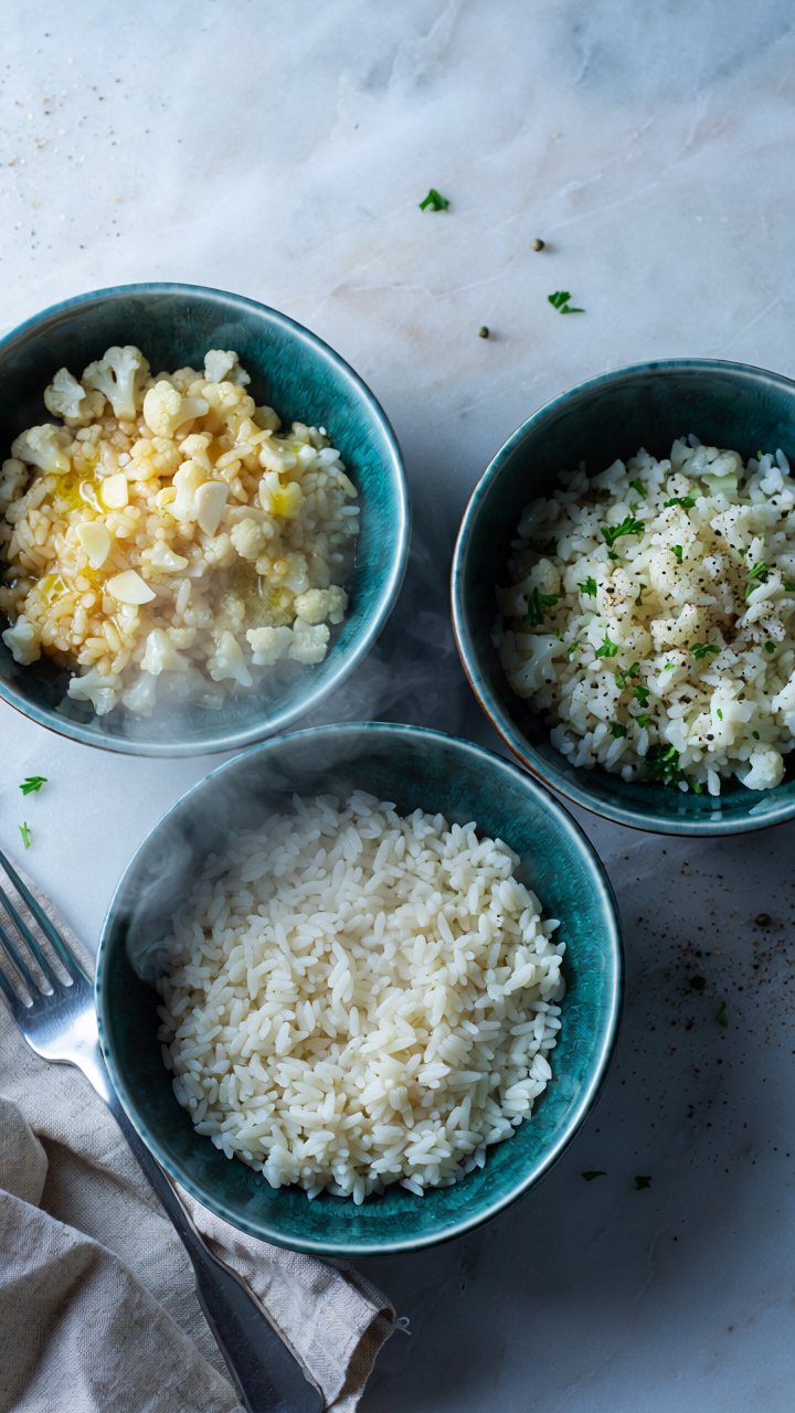 Three teal bowls of steaming cauliflower rice on a marble surface: one buttery with garlic, one with parsley and black pepper, one plain