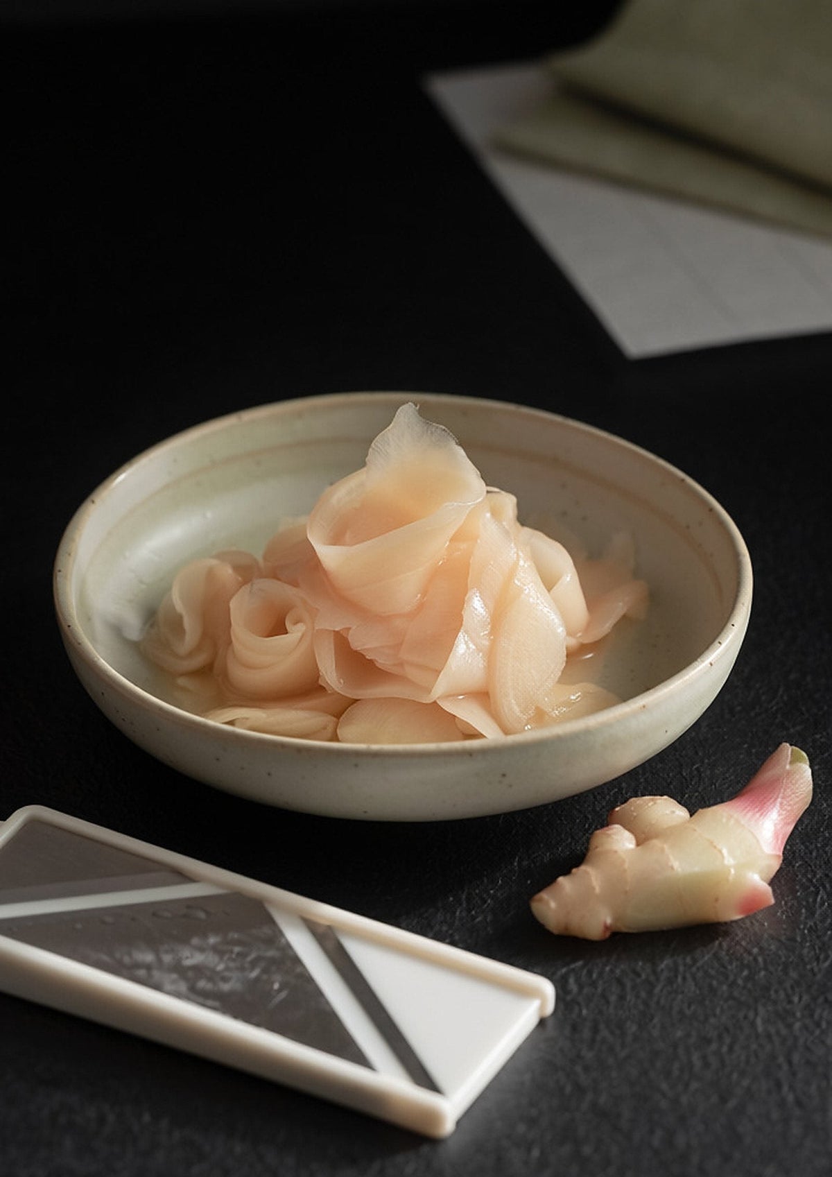 A pale ceramic dish of translucent pale pink pickled ginger slices, with a mandoline and a knob of pink-tipped young ginger beside it on a dark surface