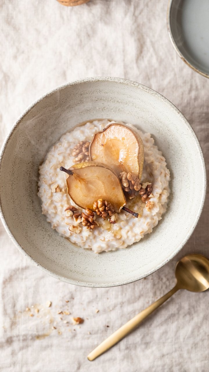 A speckled stoneware bowl of creamy oat porridge topped with roasted pear halves and toasted walnuts, on a linen cloth with a gold spoon