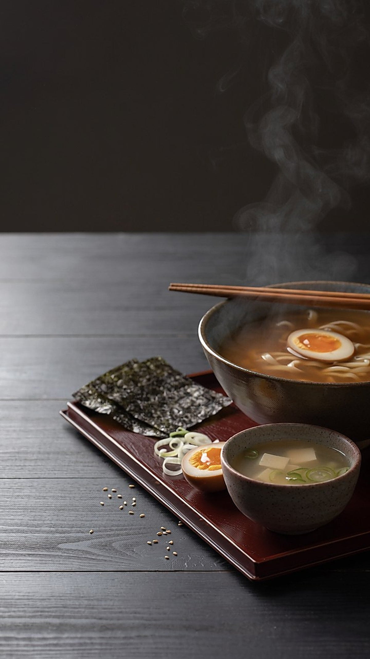 A steaming bowl of ramen with a marinated soft egg and chopsticks resting on the rim, on a lacquered tray with nori and a small bowl of miso soup