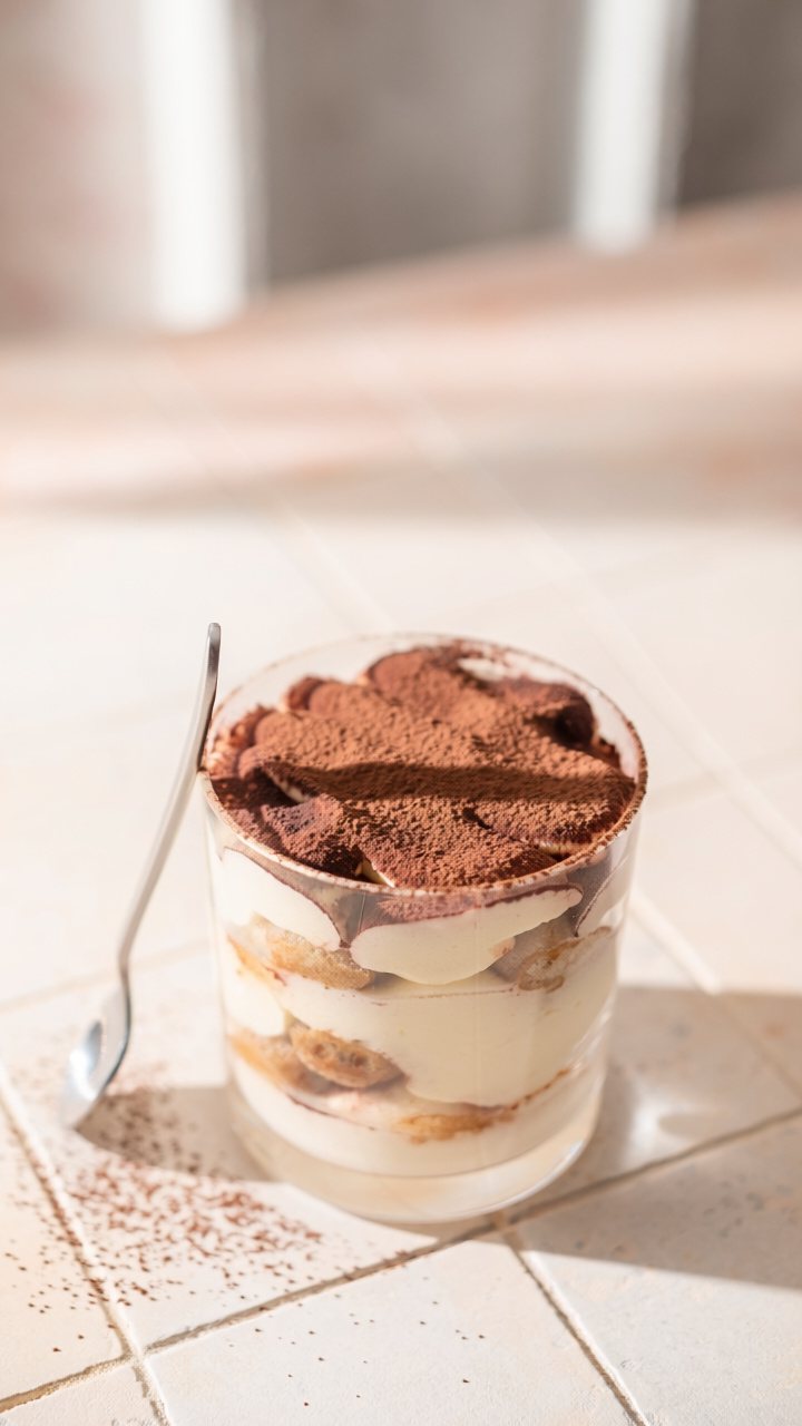 Tiramisu layered in a glass tumbler, with coffee-soaked sponge fingers, mascarpone cream and a thick dusting of cocoa, a spoon leaning beside it in the sun