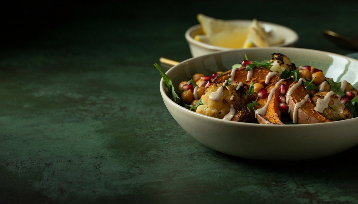 A stoneware bowl of roasted cauliflower, sweet potato and chickpeas drizzled with tahini and scattered with pomegranate seeds and herbs, on a dark green surface